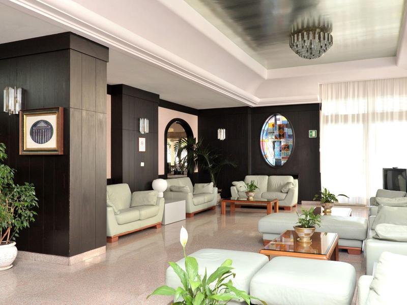 Lichte hotel lounge met witte banken, planten en grote ramen met gordijnen.