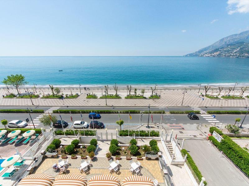 Bovenaanzicht van een strandpromenade met parkeerplaatsen, bomen en de zee op de achtergrond.