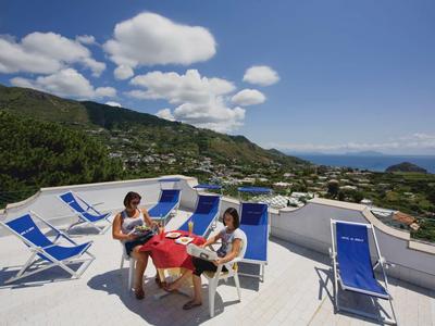 Drei Personen sitzen an einem Tisch auf einer Dachterrasse mit blauen Liegestühlen und Bergblick.