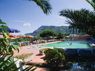 Schwimmbad mit bunten Sonnenschirmen, grünen Pflanzen und Berg im Hintergrund unter blauem Himmel.