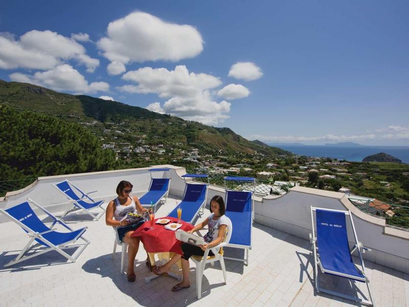 Drei Personen sitzen an einem Tisch auf einer Dachterrasse mit blauen Liegestühlen und Bergblick.