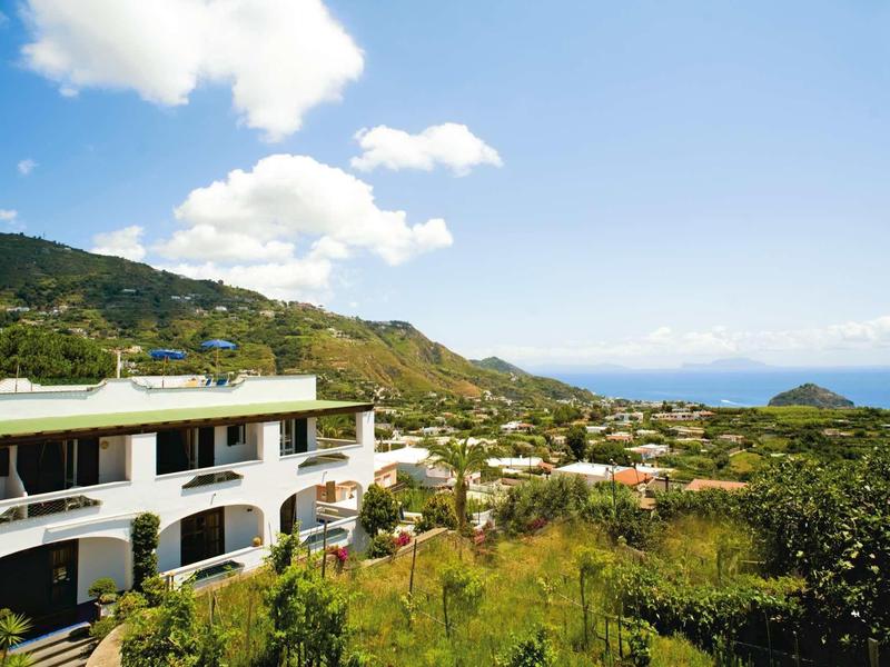 Weißes Gebäude mit Arkaden vor grünen Hügeln und weitem Meer unter blauem Himmel.