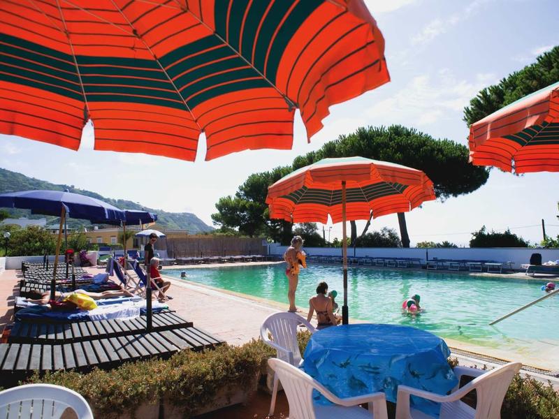 Schwimmbad mit roten Sonnenschirmen, blauem Tisch und Menschen im Wasser und am Rand.