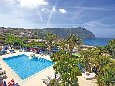 Blick auf einen blauen Pool, Palmen und Sonnenschirme vor einer Küstenlandschaft mit Hügeln und blauem Himmel.