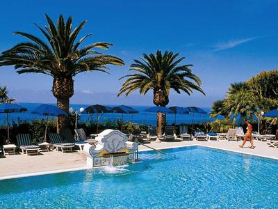 Großer blauer Pool mit weißen Liegestühlen, Palmen und Meer im Hintergrund bei Sonnenschein.