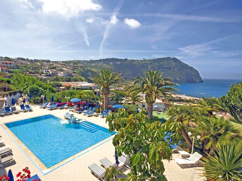 Blick auf einen blauen Pool, Palmen und Sonnenschirme vor einer Küstenlandschaft mit Hügeln und blauem Himmel.