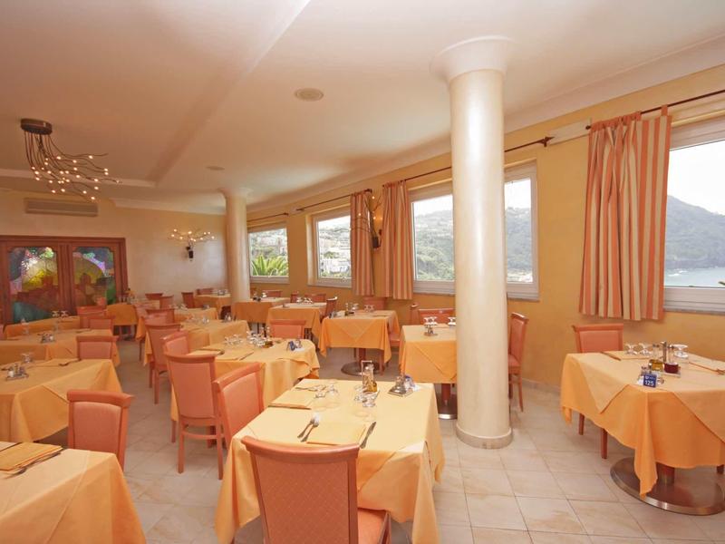 Restaurant mit rechteckigen Tischen, orangenen Tischdecken, Holzstühlen und großen Fenstern mit Vorhängen.