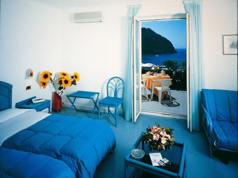 Helles Zimmer mit blauer Einrichtung, Sonnenblumen, offenem Balkon und Meerblick.