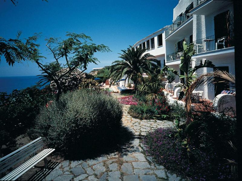 Haus mit Terrasse und Pflanzen an einem sonnigen Küstenweg mit Blick aufs Meer.