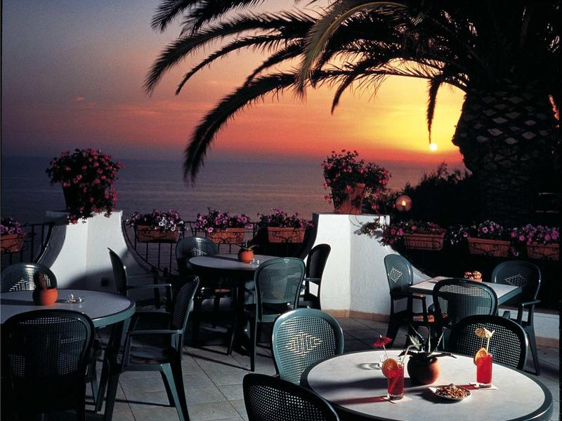 Terrassenbereich mit Tischen, Stühlen und Palmen vor einem roten Sonnenuntergang am Meer.