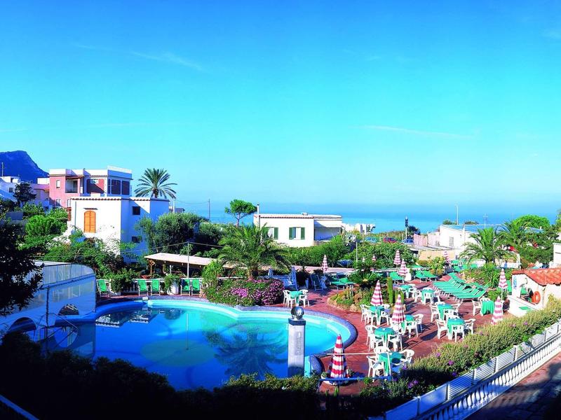 Complesso alberghiero con piscina, lettini e vista mare sotto cielo sereno.