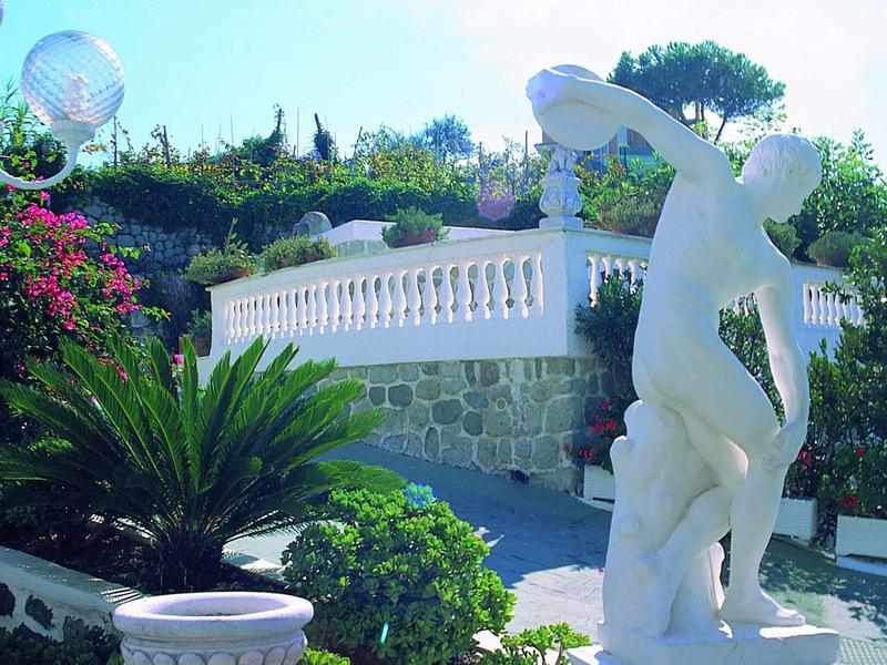 Fontana in pietra con statua bianca accanto a piante verdi rigogliose e architettura mediterranea.