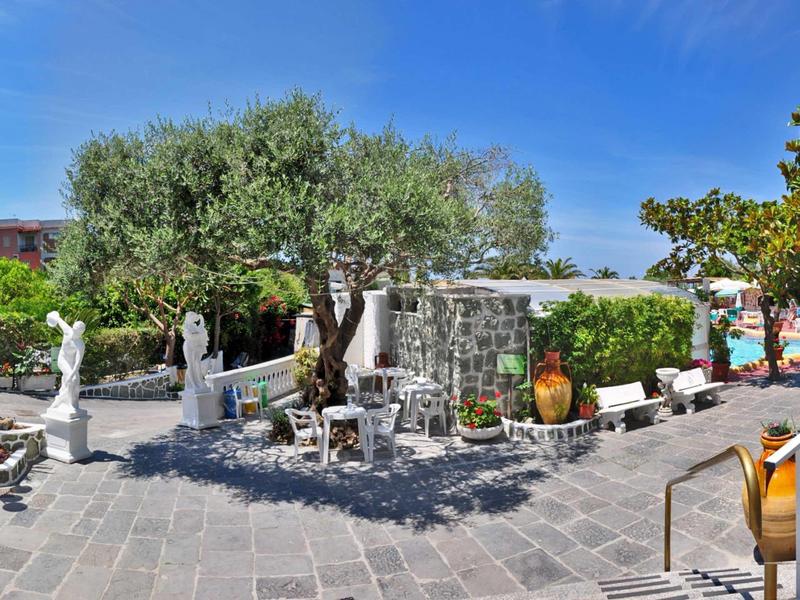 Area esterna con terrazza in pietra, mobili bianchi e ulivi sotto un cielo blu