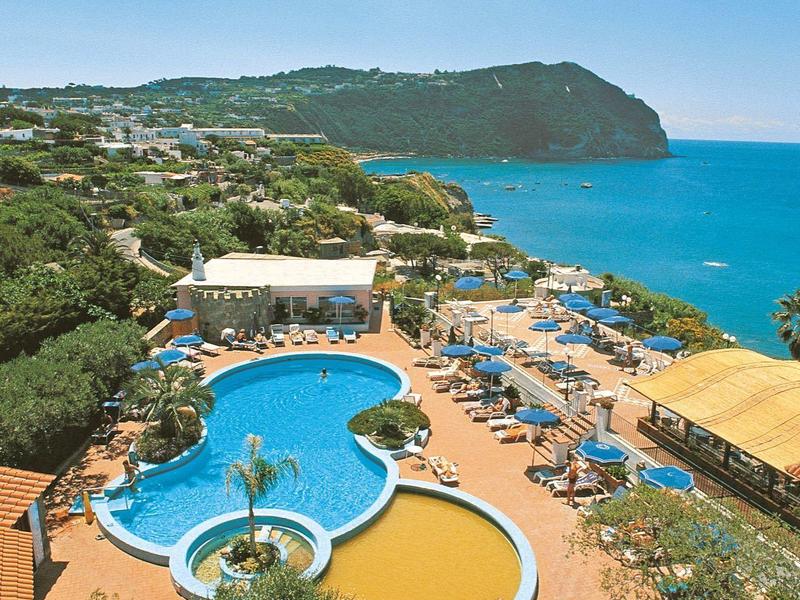 Meerdere zwembaden met ligstoelen aan een kustlijn met blauwe zee en bewolkte lucht.