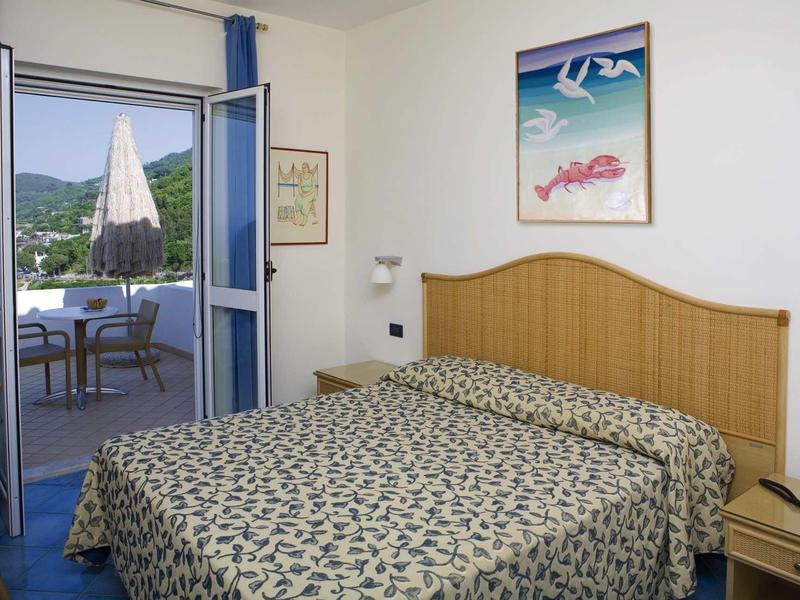 Bright hotel room with double bed and access to balcony with table and chairs.