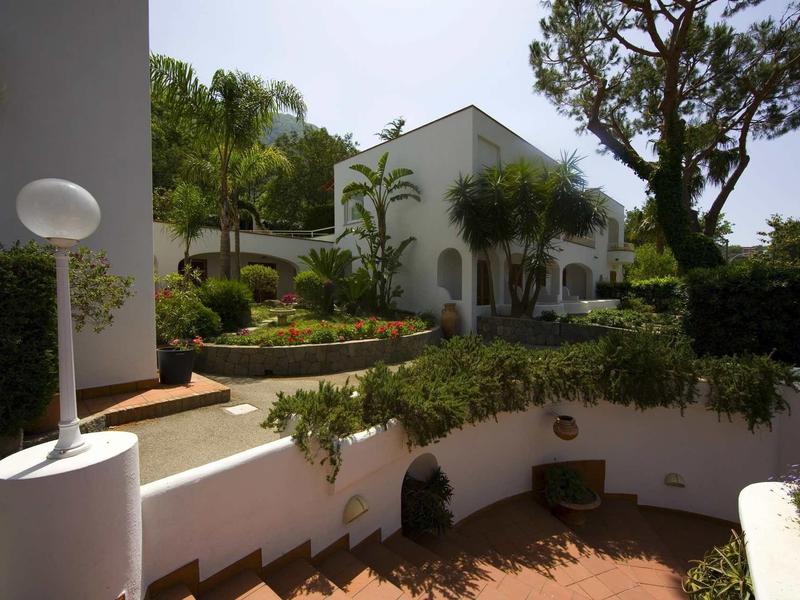 House with garden, white walls, and paved path surrounded by trees and bushes.