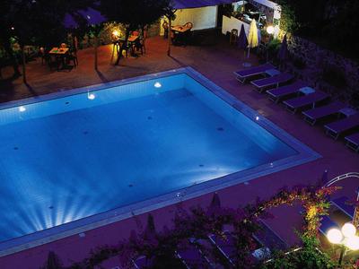 Beleuchteter rechteckiger Pool mit Liegestühlen und Pflanzen in einer Hotelanlage bei Nacht.
