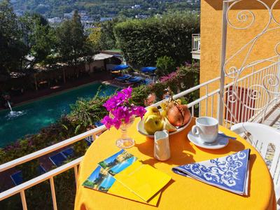 Bunter Frühstückstisch mit Tasse, Obst und Serviette auf Balkon mit Blick auf Pool und Garten.
