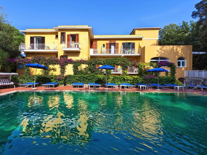 Großes gelbes Gebäude mit mehreren Balkonen, Sonnenschirmen und Liegestühlen neben einem Pool.