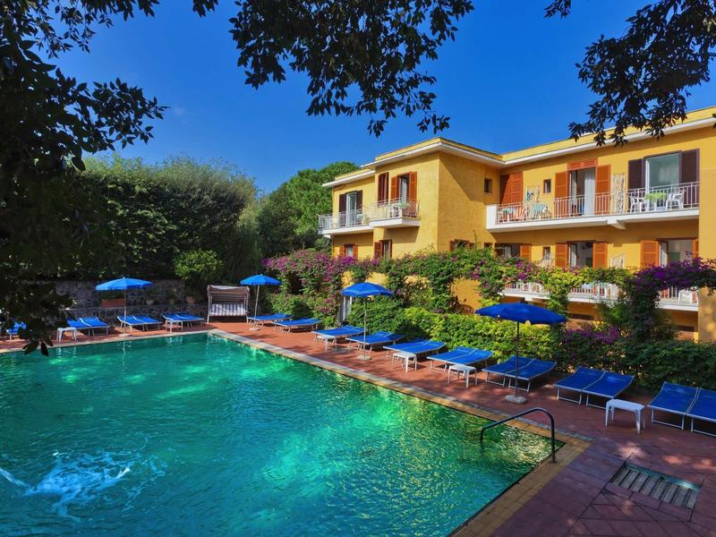 Schwimmbad mit blauem Wasser, umgeben von blauen Liegestühlen und gelbem Gebäude unter blauem Himmel.