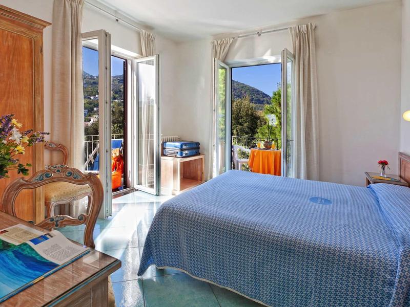 Helles Schlafzimmer mit blauem Bett, offenen Fenstern und Blick auf Berglandschaft.