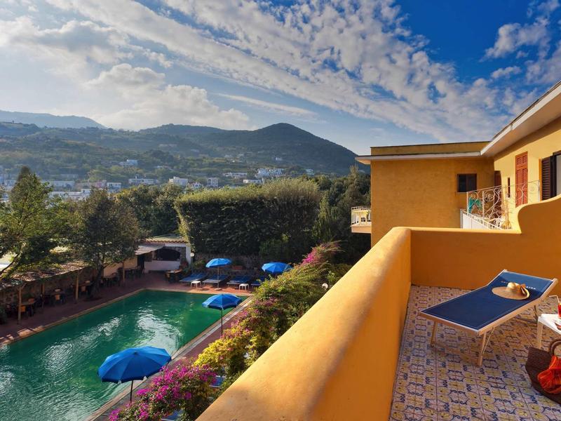 Blick von Balkon auf blauen Pool, grüne Bäume und hügelige Landschaft unter blauem Himmel.