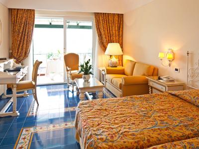 Světly hotelový pokoj s manželskou postelí, pohovkou, pracovním stolem a přístupem na balkon s výhledem ven.