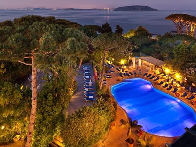 Beleuchteter Hotelpool umgeben von Sonnenliegen und mediterranen Bäumen bei Dämmerung mit Meerblick.