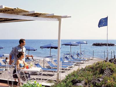 Zwei Personen gehen an einer Strandpromenade mit Liegestühlen und blauen Sonnenschirmen am Meer vorbei.