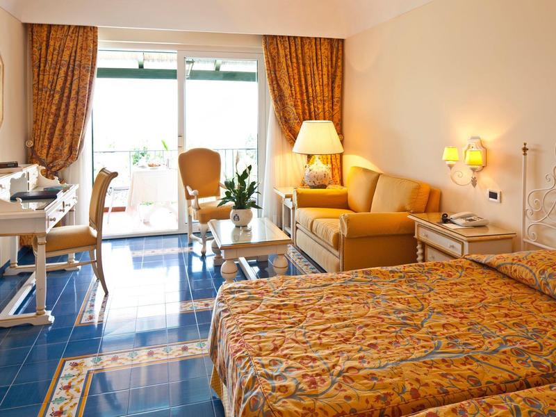 Světly hotelový pokoj s manželskou postelí, pohovkou, pracovním stolem a přístupem na balkon s výhledem ven.