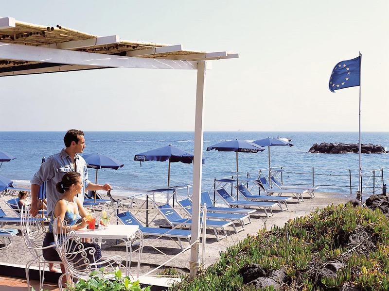 Dvě osoby užívající si výhled na lehátka u moře pod jasně modrou oblohou.