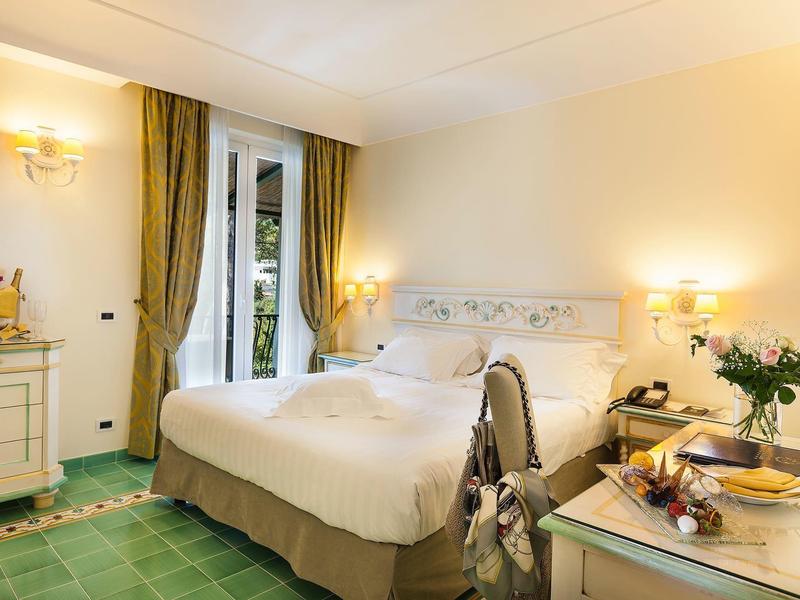 Pohodlný hotelový pokoj s velkou postelí, zelenými závěsy a dlaždicovou podlahou.