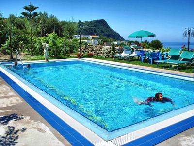 Ein rechteckiger Pool mit klar blauem Wasser, umgeben von Liegestühlen und Sonnenschirmen, mit Bergen im Hintergrund.