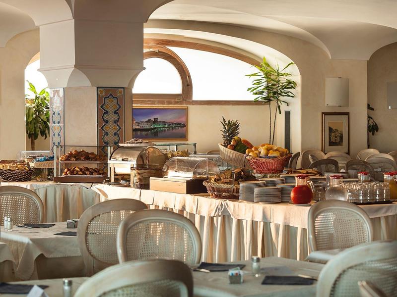 Helles Restaurant mit rundem Buffet-Tisch, gedeckten Tischen und großen Fensterbögen.