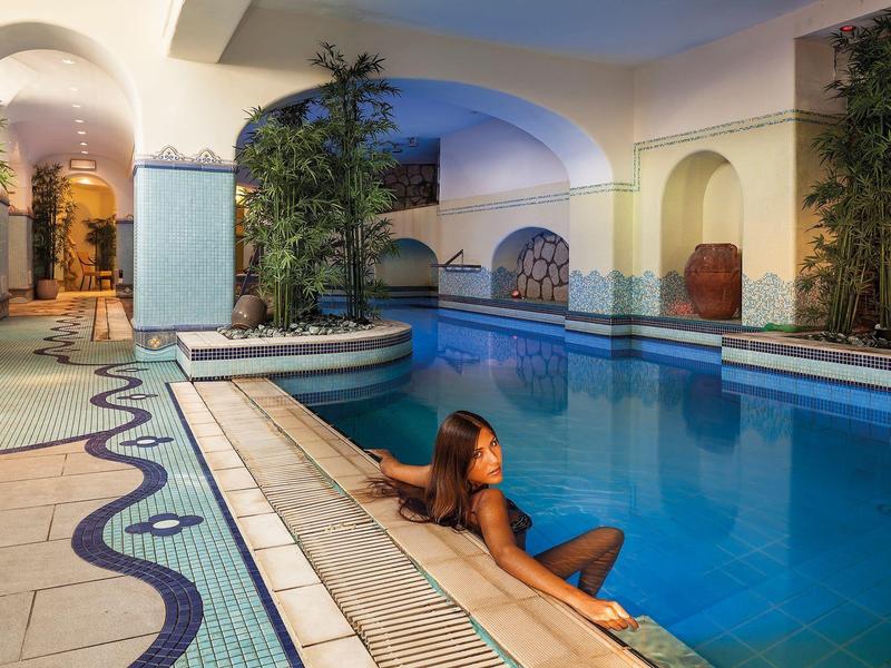 Frau sitzt entspannt am Rand eines beleuchteten Hallenbads mit weiß-blauem Fliesenmuster und Palmen.