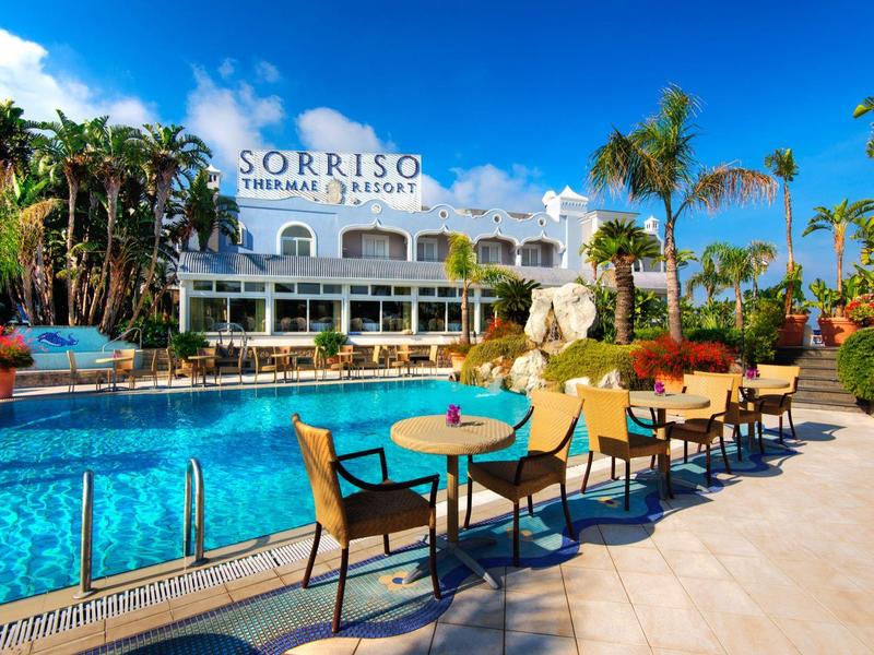 Großer Pool mit Sonnenterrasse, Tischen und Stühlen vor einem Hotel mit Palmen und blauem Himmel.