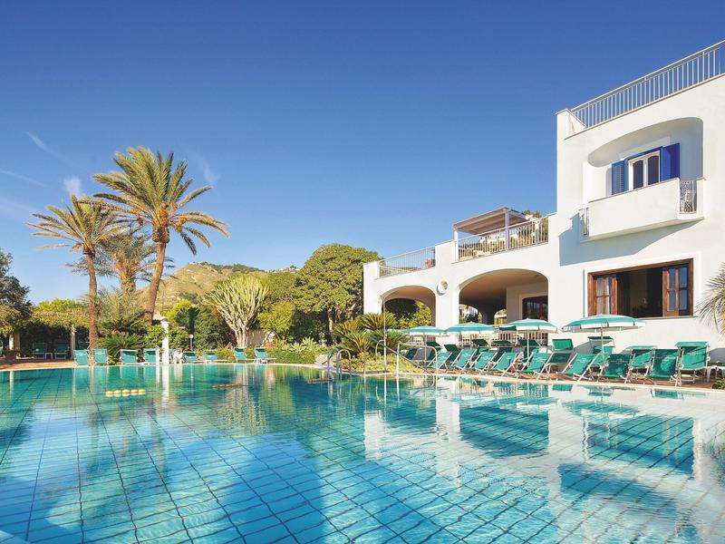 Duży basen na zewnątrz przed białym budynkiem hotelu z parasolami i palmami.