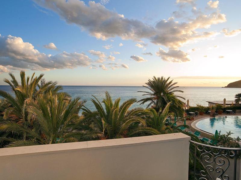 Widok na basen hotelowy z palmami i morzem pod częściowo zachmurzonym niebem podczas zachodu słońca.