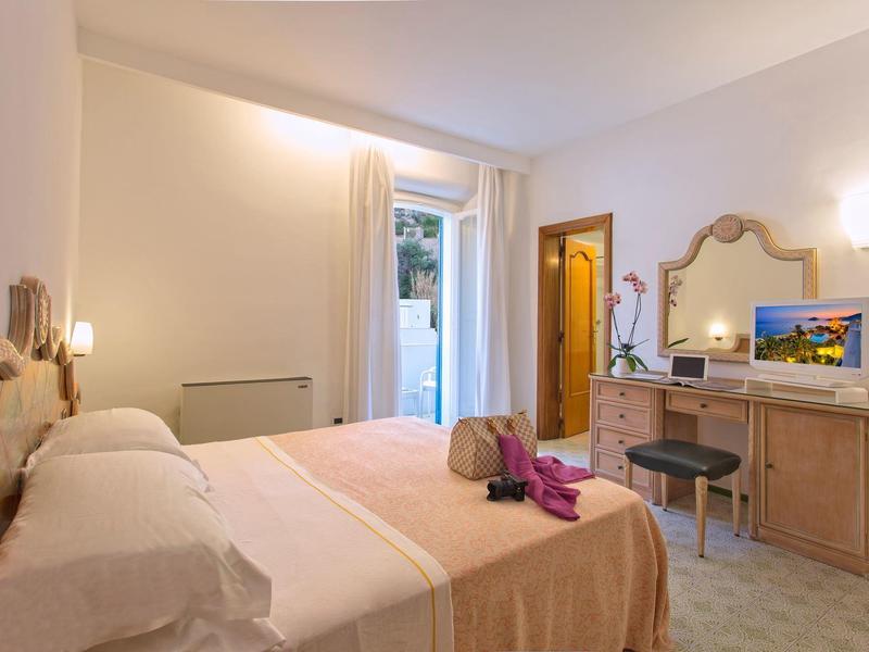 Jasny pokój hotelowy z łóżkiem podwójnym, biurkiem, krzesłem i balkonem z widokiem na zewnątrz.