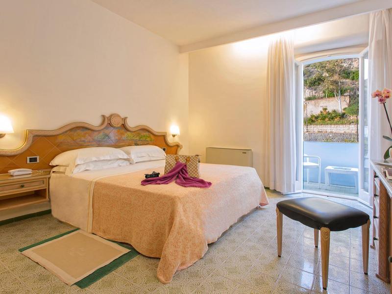 Jasny pokój hotelowy z podwójnym łóżkiem, stolikami nocnymi, widokiem z okna i eleganckim krzesłem.