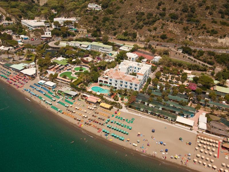 Widok z powietrza na hotel z plażą i leżakami nad morzem, otoczony wzgórzami.