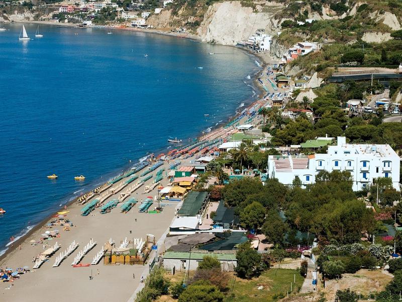 Wybrzeże z piaszczystą plażą, niebieską wodą, leżakami i białymi budynkami wśród drzew.