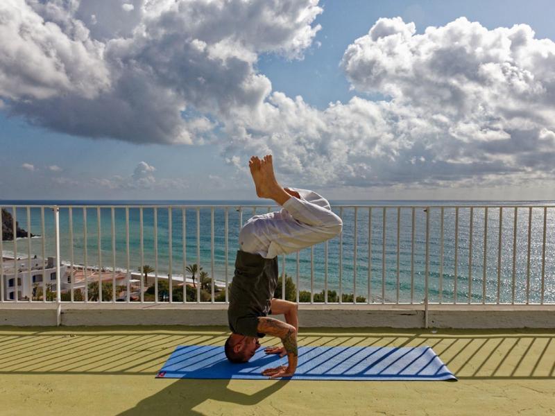 Osoba wykonująca stanie na głowie na macie do jogi na balkonie z widokiem na morze w słoneczny dzień.