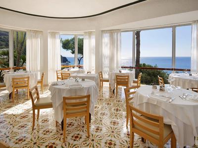 Světla restaurace s velkými okny, bílými ubrusy a výhledem na moře.