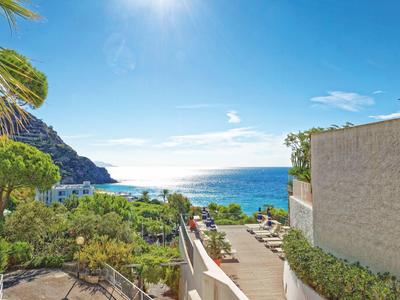 Výhled na moře z terasy s židlemi a zelenými rostlinami pod slunečnou oblohou.