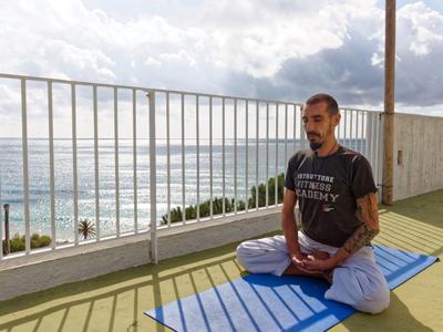 Uomo medita a gambe incrociate su un tappetino da yoga su una terrazza con vista mare.