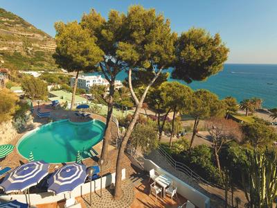Piscina dell'hotel con ombrelloni blu, pini e vista mare con cielo sereno.
