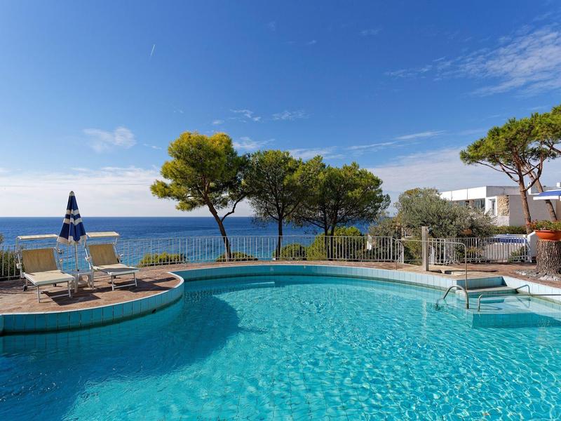 Kruhový bazén s výhledem na moře a lehátky pod modrou oblohou.