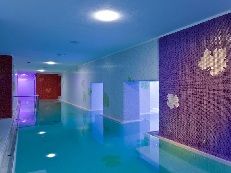 Moderní krytý bazén s modrozelenou vodou a fialovou stěnovou dekorací v hotelovém spa.