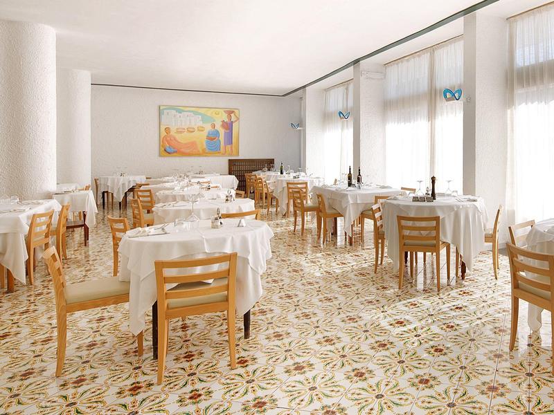 Světlá, moderní jídelna s kulatými stoly, dřevěnými židlemi, dekorativní podlahou a velkými okny.